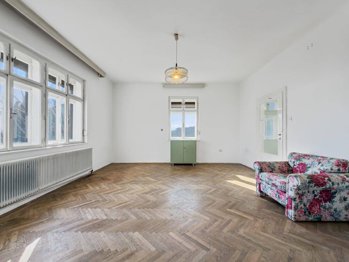 Geräumiges, unmöbliertes Zimmer mit Fischgrätparkett und floralem Sessel, vielseitig nutzbar.