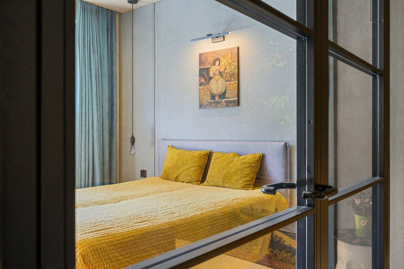 Schlafzimmer mit gelber Bettwäsche, durch eine moderne Glaswand abgetrennt.