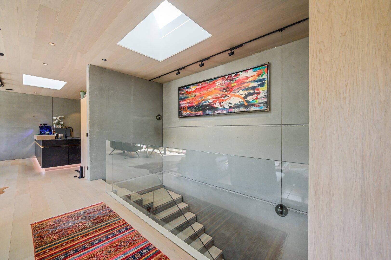 Heller Flur mit Treppe, Glasgeländer und Kunstwerk an der Wand.
