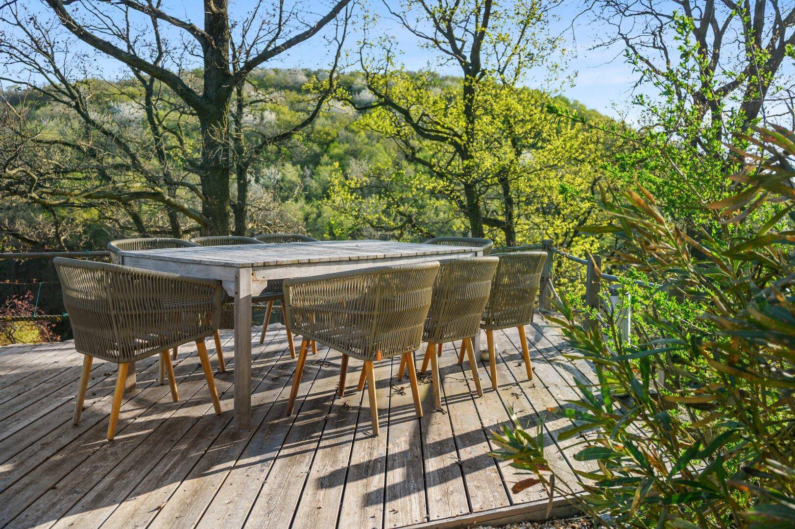 Holzdeck mit großem Esstisch und Stühlen, umgeben von Bäumen.