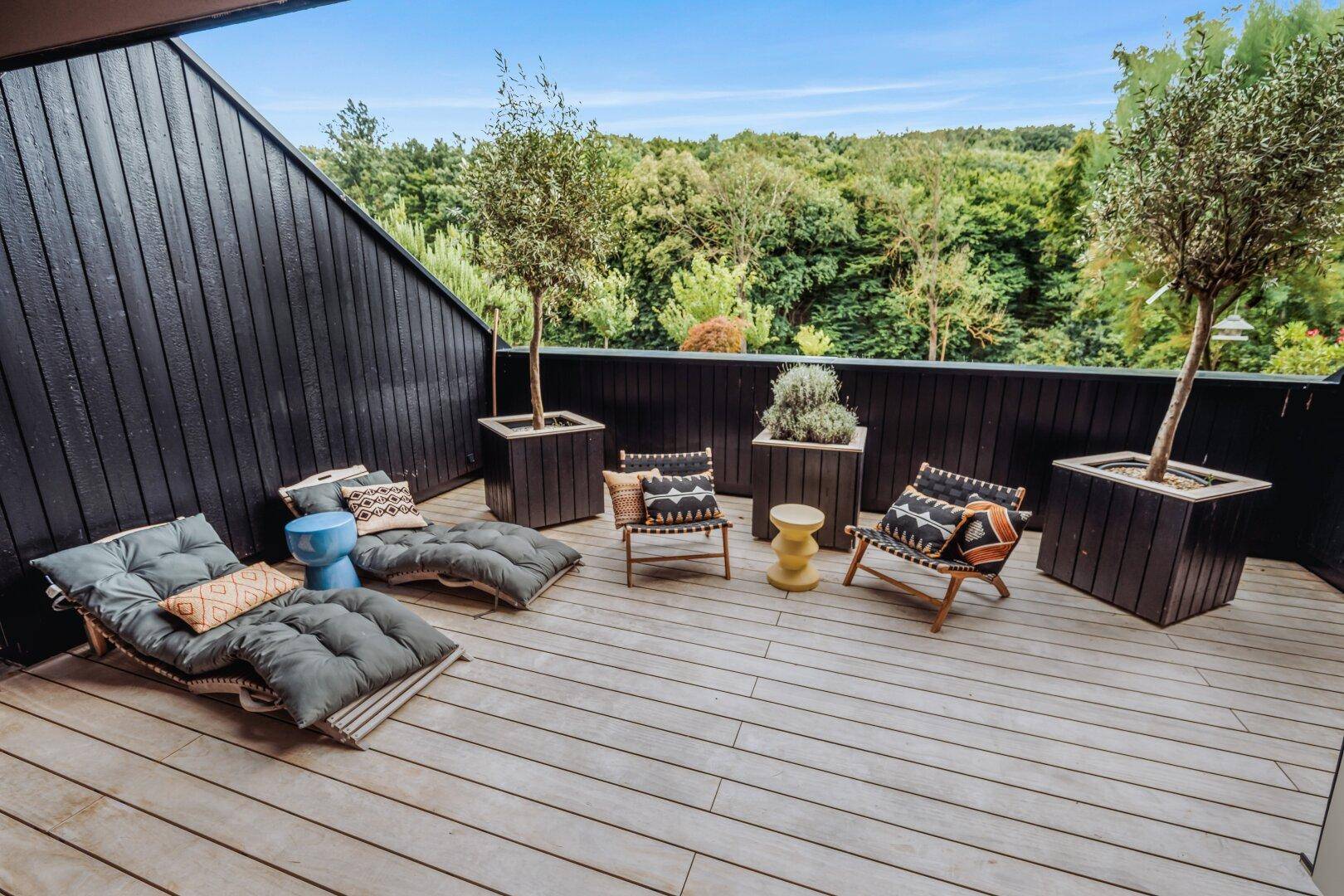 Große Holzterrasse mit Liegestühlen und Pflanzen, umgeben von grüner Natur.