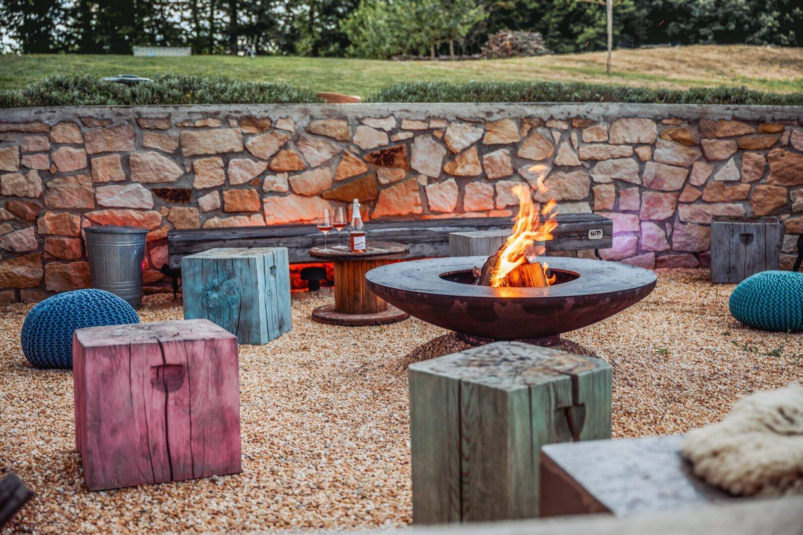 Gemütliche Feuerstelle im Garten mit bunten Sitzgelegenheiten und Steinmauer.
