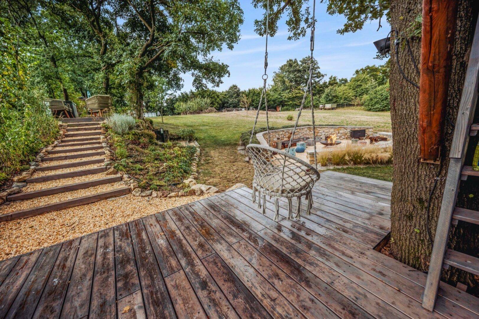 Holzdeck mit Schaukel und Blick auf die Feuerstelle im weitläufigen Garten.