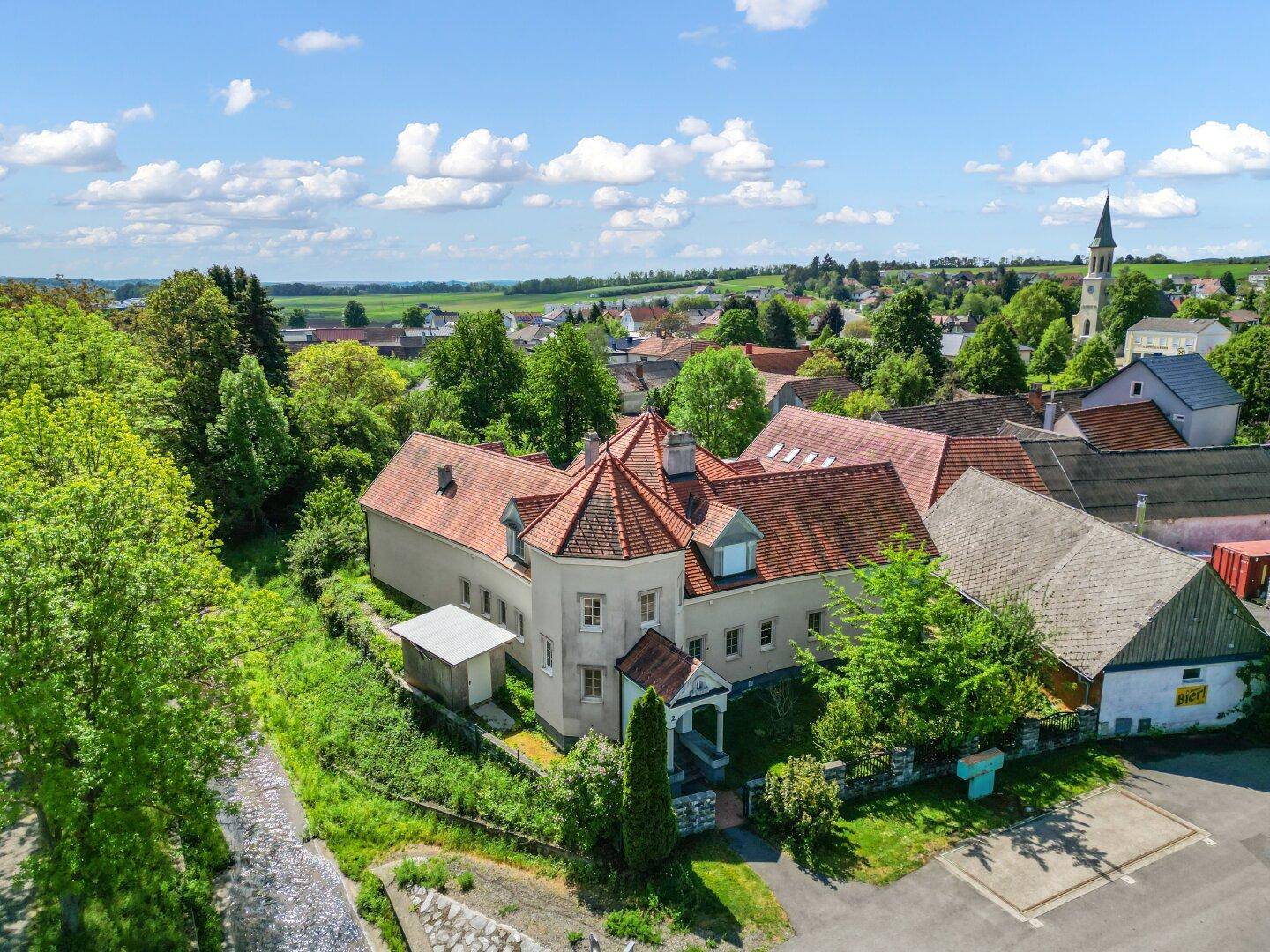Luftaufnahme eines Anwesens mit markantem roten Ziegeldach, umgeben von viel Grün und einem Dorf.