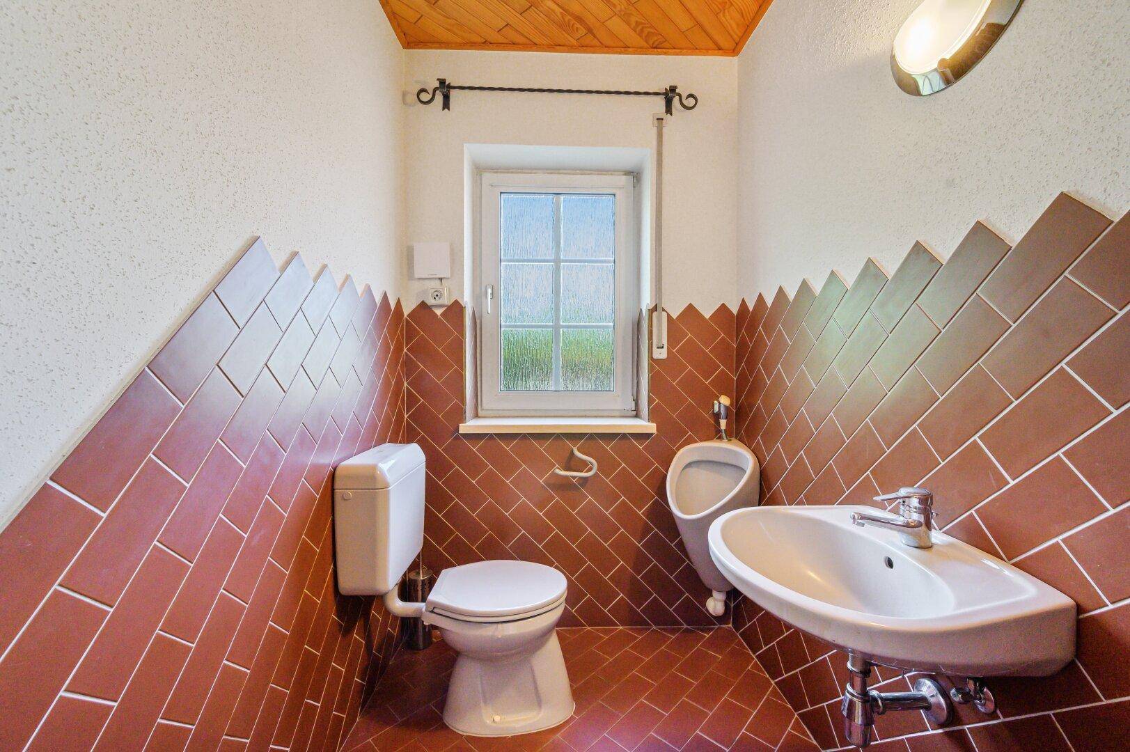 Gästetoilette mit braunen Fliesen, Urinal, Waschbecken und Fenster.