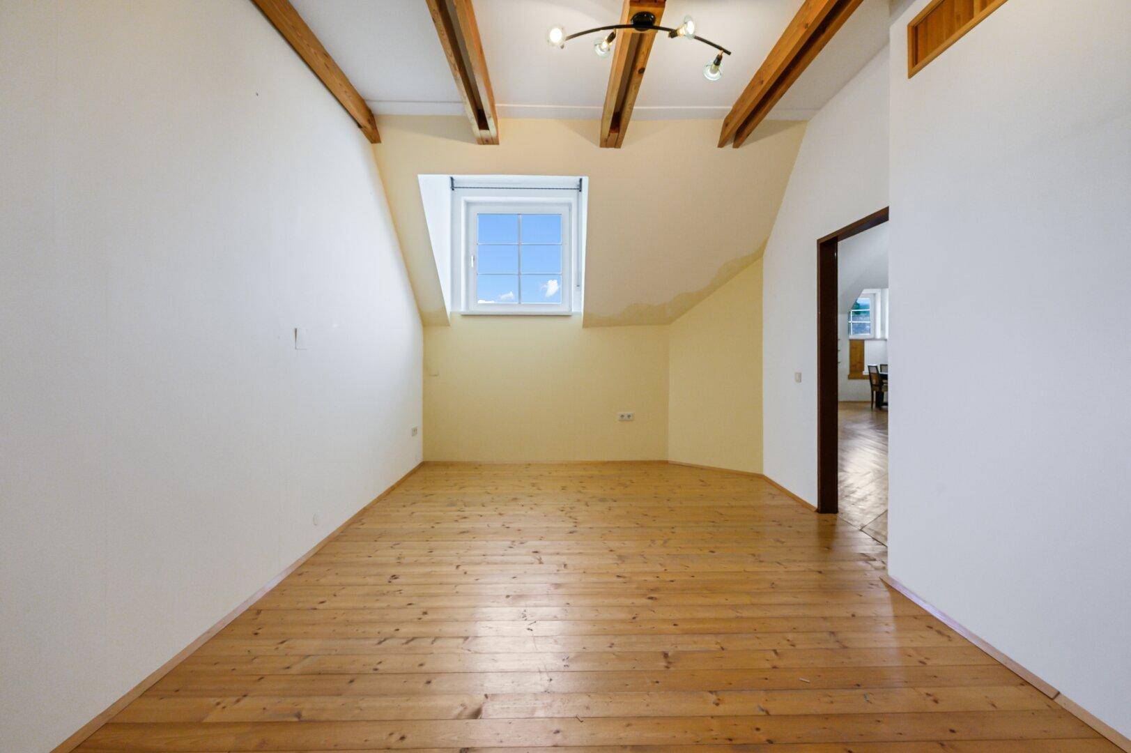 Leeres Zimmer mit Holzboden, schräger Decke, sichtbaren Balken und einem Dachfenster.
