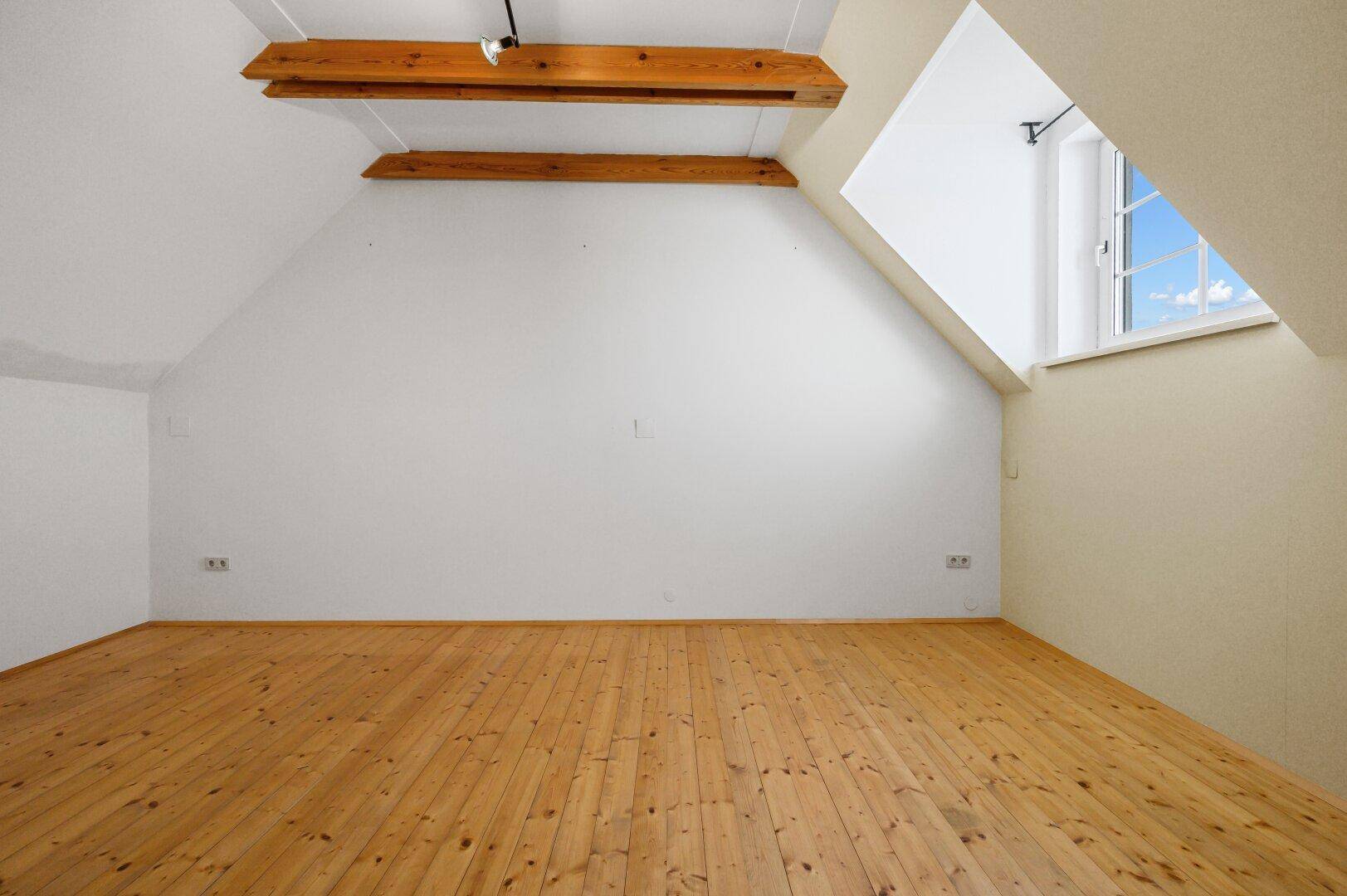 Geräumiges Zimmer mit Holzboden, schräger Decke und sichtbaren Holzbalken.