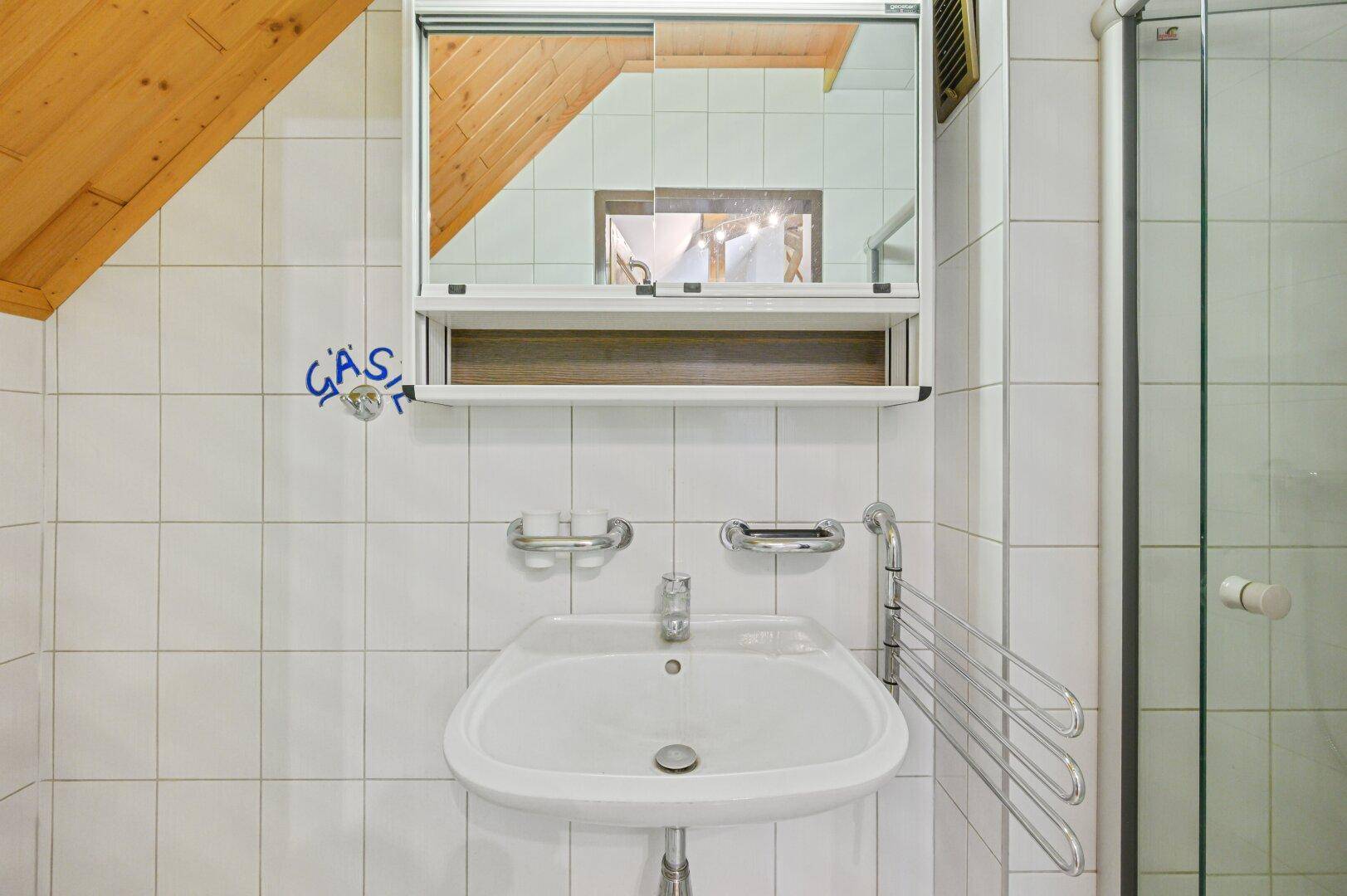Badezimmer mit weißen Fliesen, Einzelwaschbecken, Spiegelschrank und Duschkabine.