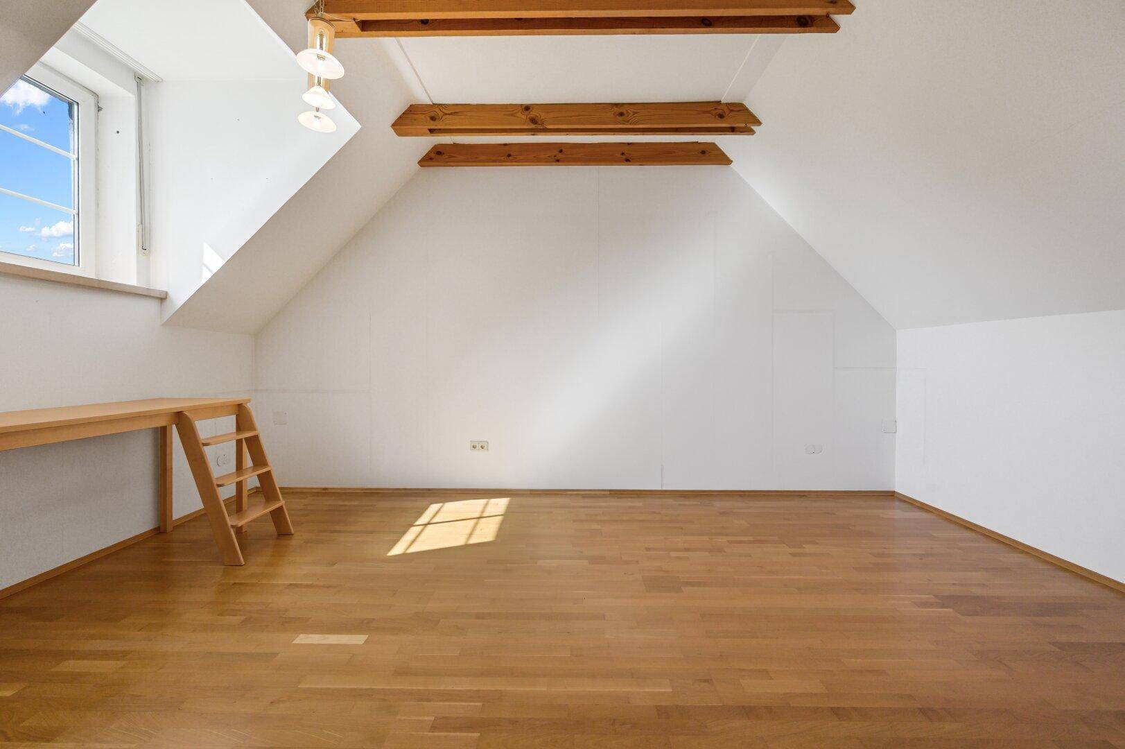 Helles, leeres Zimmer mit Holzboden, schräger Decke und sichtbaren Holzbalken.