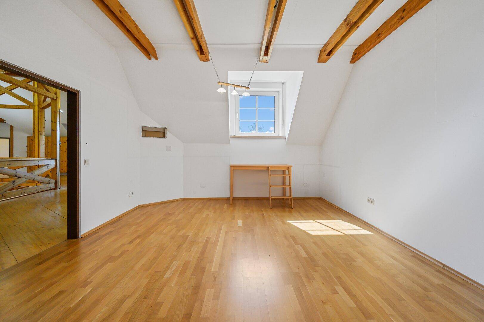 Geräumiges Zimmer mit Holzboden, schräger Decke und Blick in den offenen Wohnbereich.