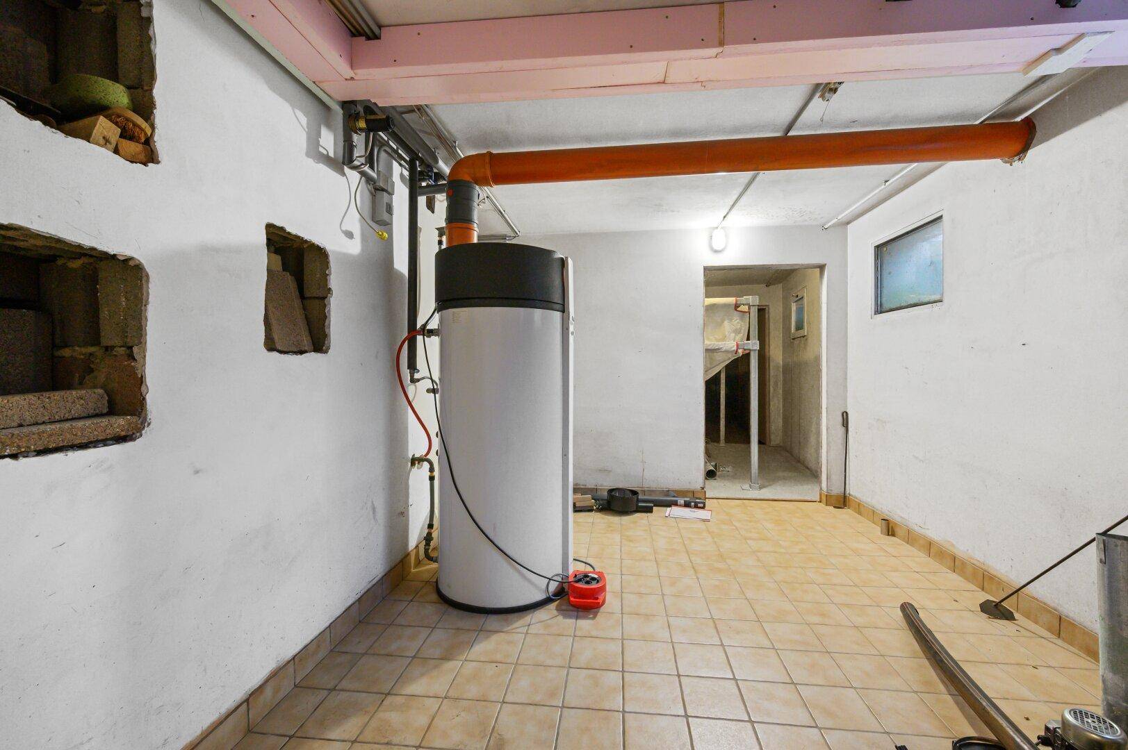 Kellerraum mit Warmwasserbereiter, sichtbaren Rohren und Fliesenboden.