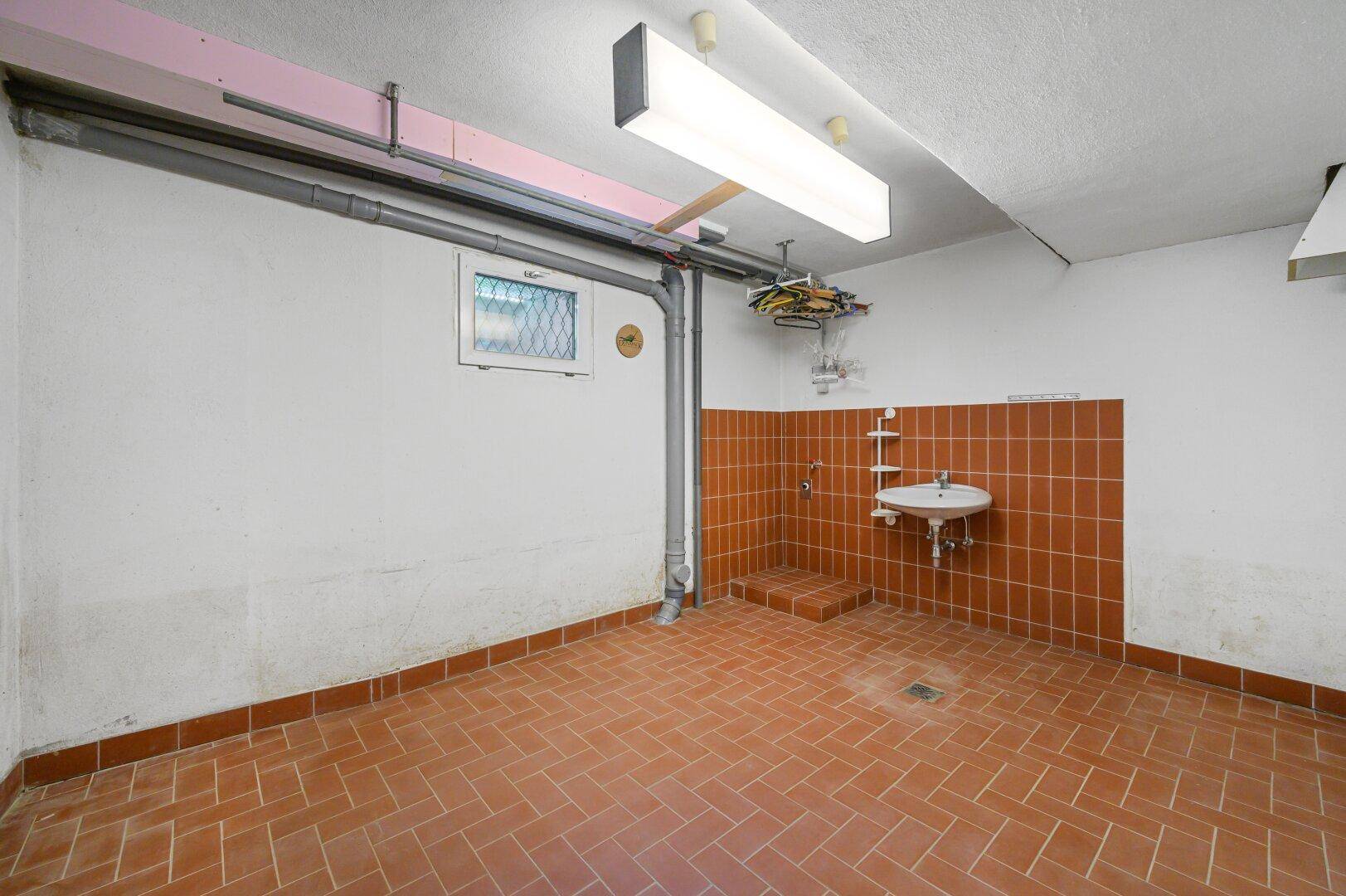 Keller-Hauswirtschaftsraum mit Terrakotta-Fliesen, kleinem Waschbecken und sichtbaren Rohren.