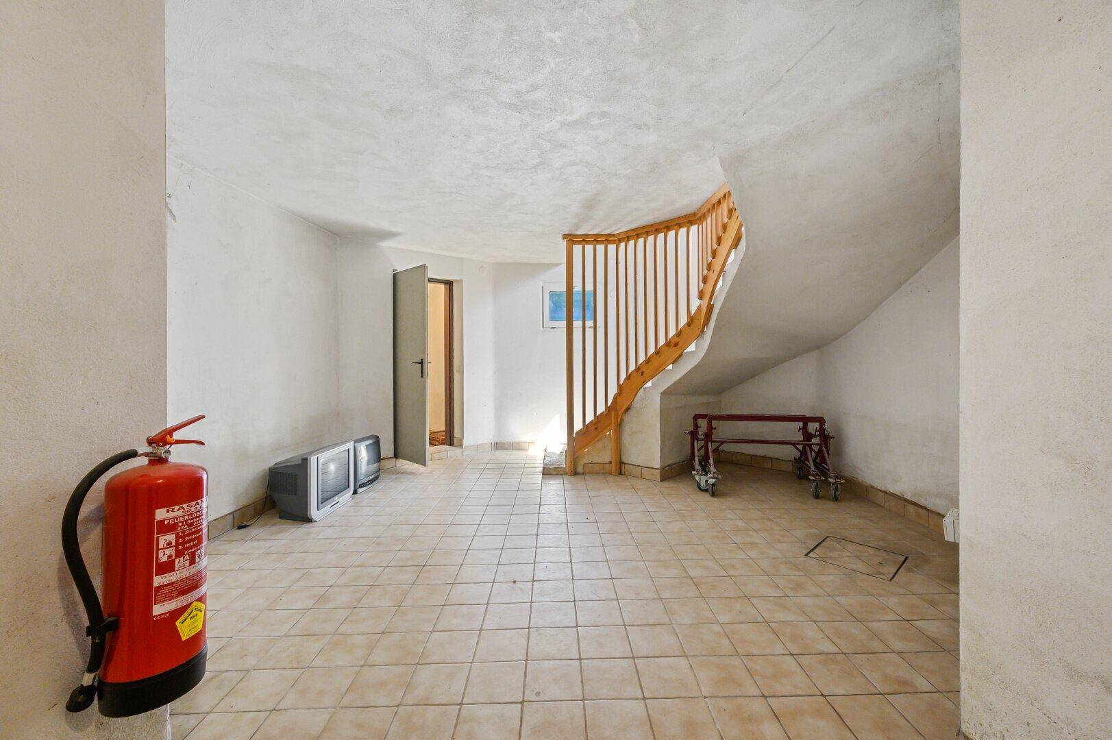 Kellerraum mit Fliesenboden, Feuerlöscher und Holztreppe, dient als Abstellfläche.