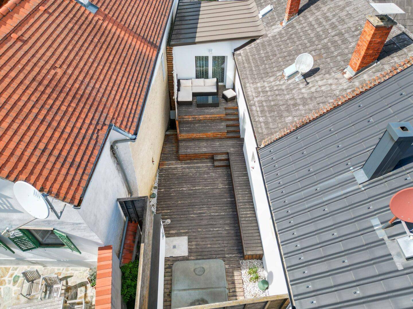 Luftaufnahme einer schmalen Holzterrasse mit Sitzbereich und Whirlpool zwischen Gebäuden.