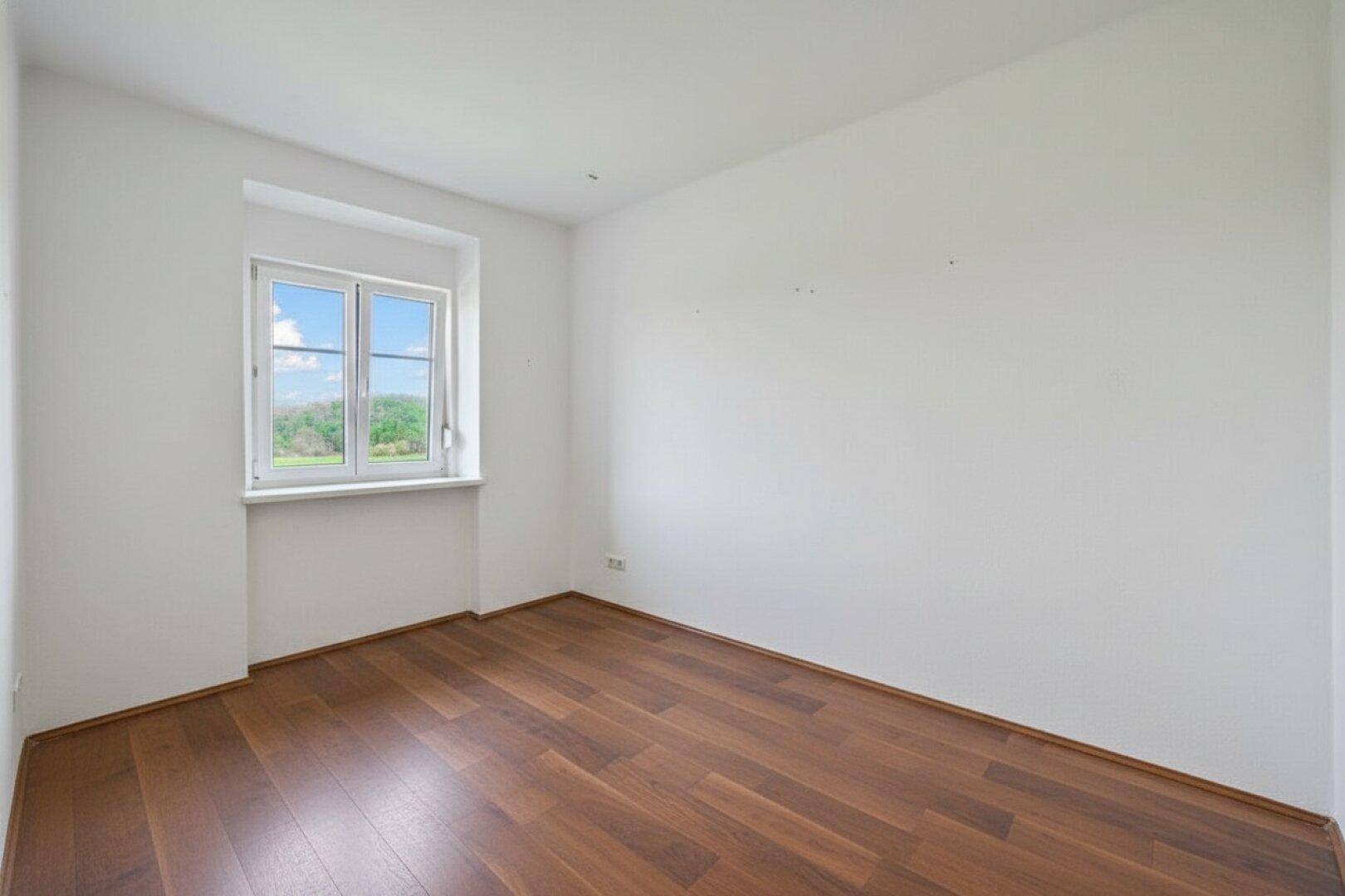 Leeres Schlafzimmer mit Holzboden und einem Fenster, das viel Licht hereinlässt.