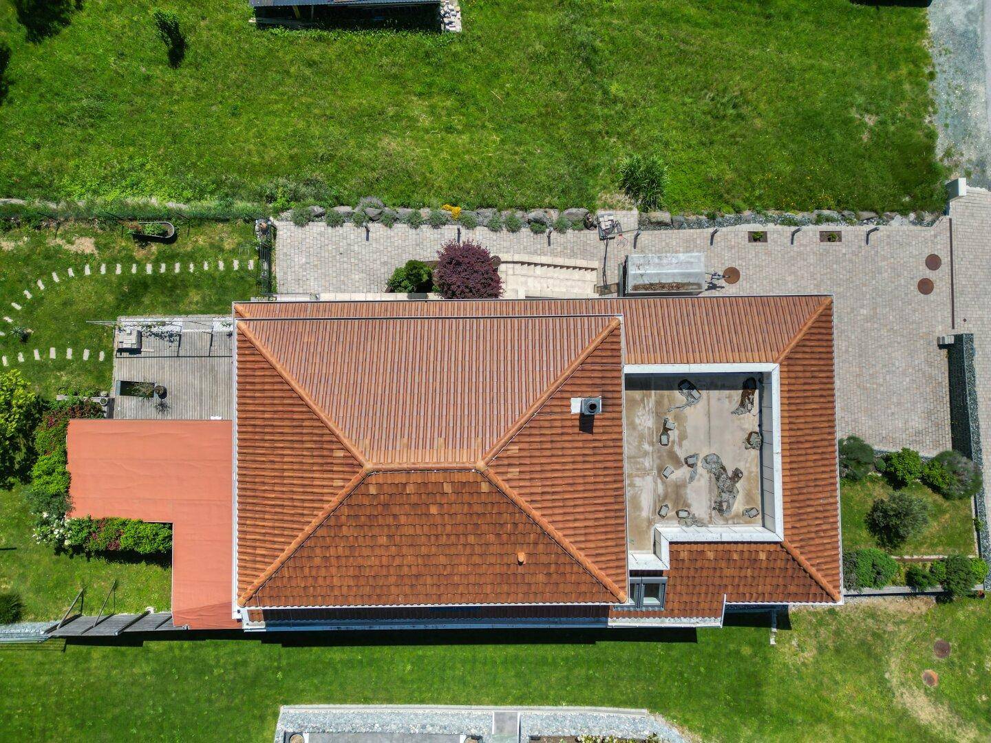 Luftaufnahme des Hausdaches mit komplexer Ziegelstruktur und einem Flachdachbereich, umgeben von Grünflächen.