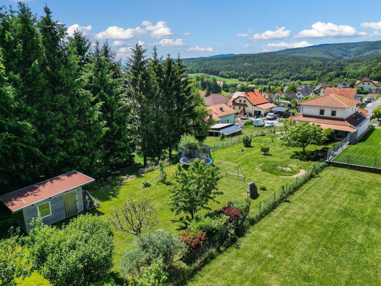Luftaufnahme eines weitläufigen Grundstücks mit Haus, großem Rasen, Bäumen, Trampolin und Gartenhaus.