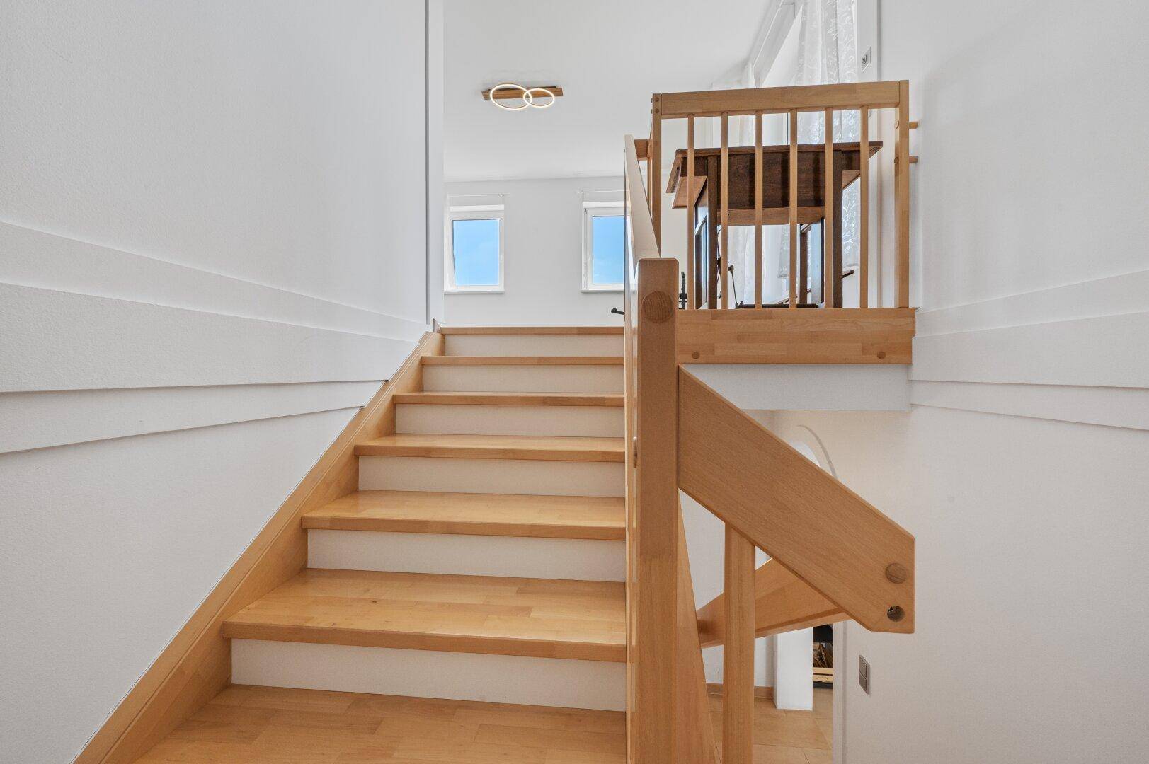 Holztreppe führt zu einem hellen Treppenabsatz mit kleinen Fenstern und einem Holztisch.