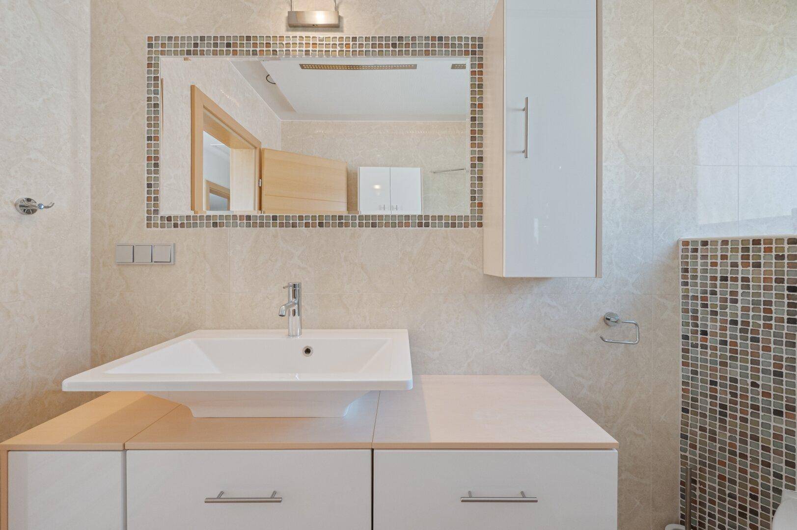 Badezimmer mit Einzelwaschbecken, großem Spiegel mit Mosaikrahmen und einem weißen Wandschrank.