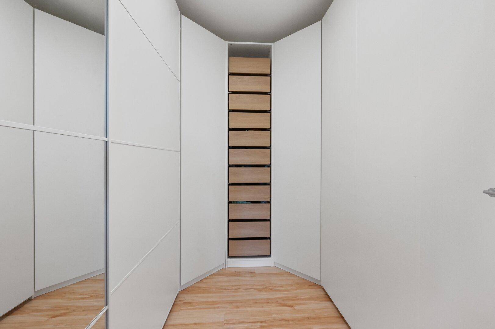 Begehbarer Kleiderschrank mit weißen Einbauschränken und einem zentralen Regal mit Schubladen.