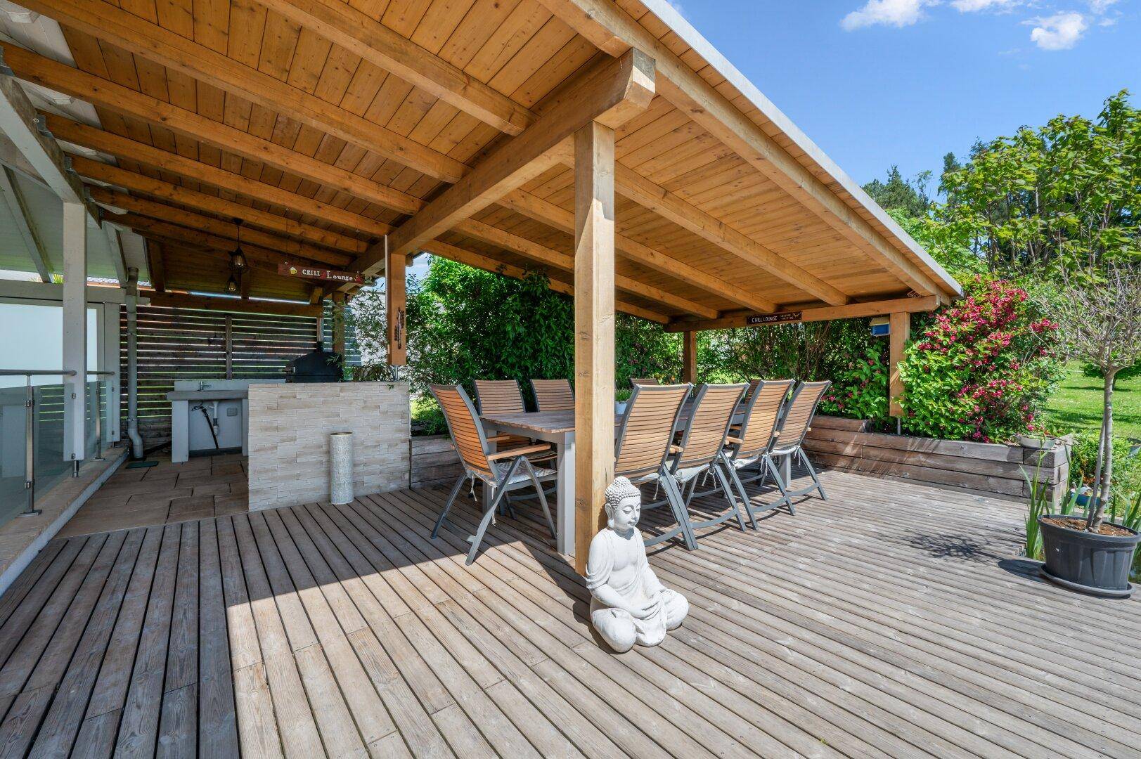 Geräumige überdachte Holzterrasse mit großem Esstisch, Buddha-Statue und integriertem Grillbereich.