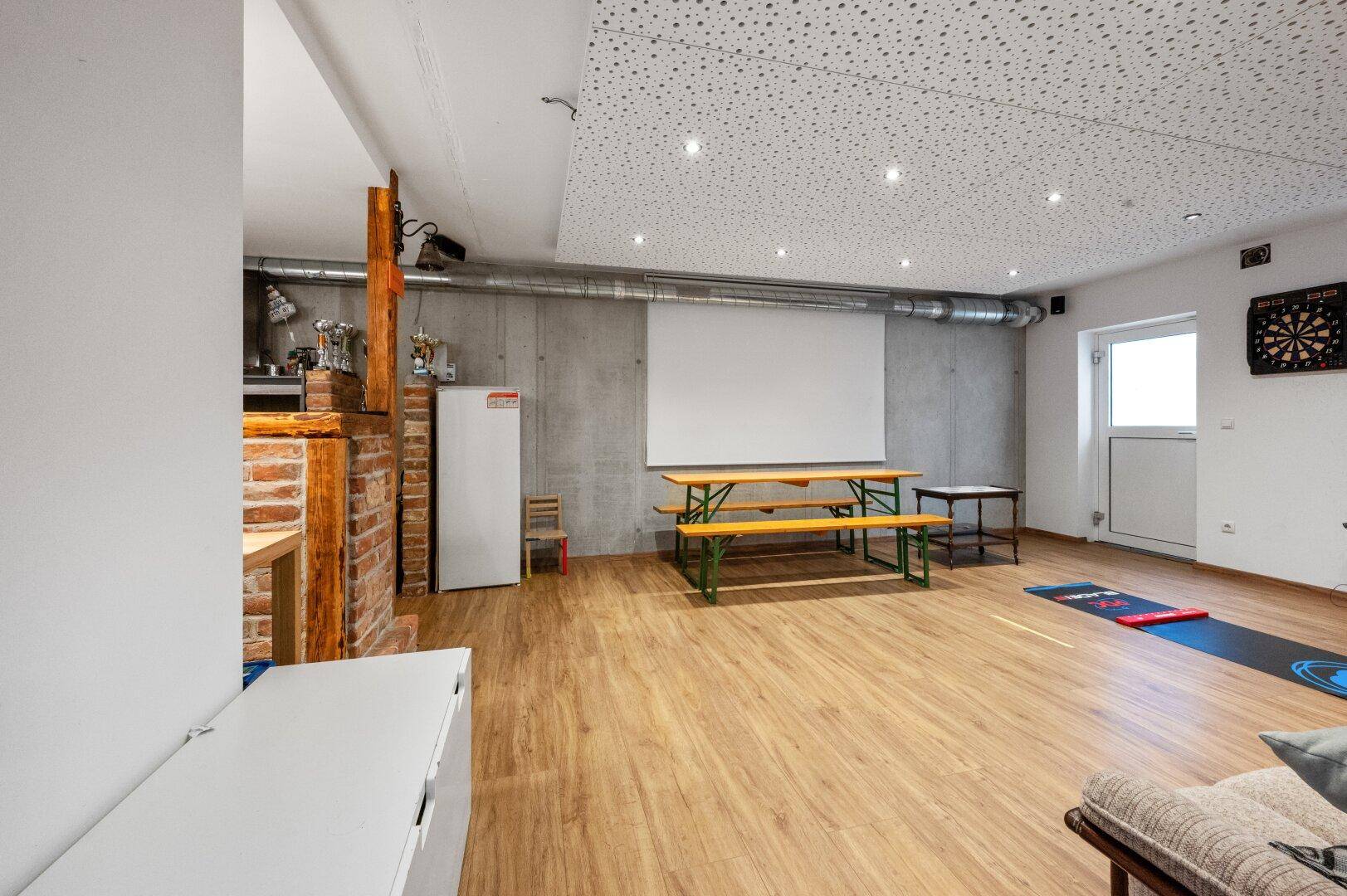 Kellerraum mit Beton- und Ziegelwänden, Laminatboden, Leinwand und Dartscheibe, ideal für Freizeitaktivitäten.