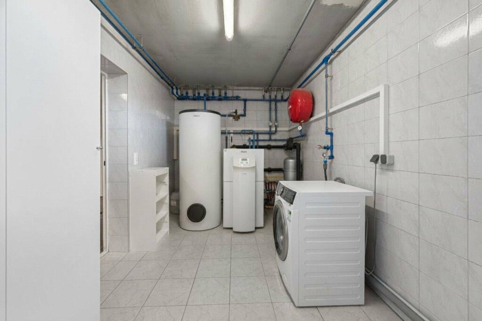 Hauswirtschaftsraum im Keller mit Waschmaschine, Warmwasserspeicher und sichtbaren Rohrleitungen.