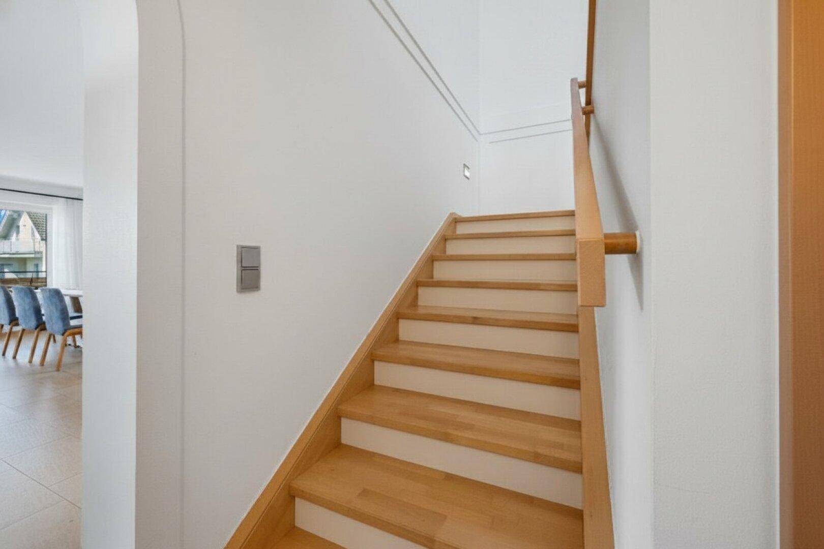 Heller Treppenaufgang mit Holzstufen und weiß gestrichenen Wänden, führt in die obere Etage.