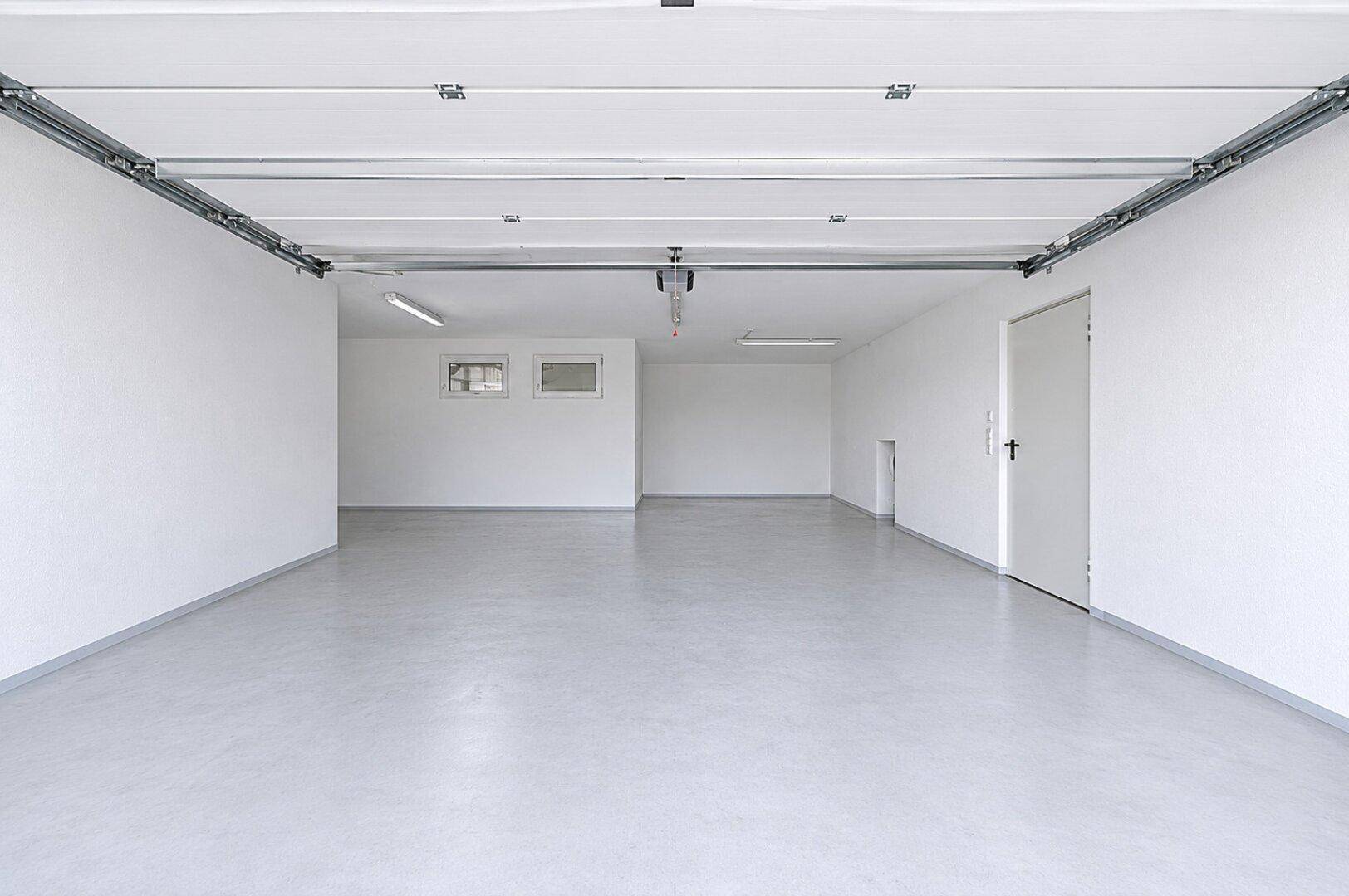 Geräumige, leere Garage mit weißen Wänden, Betonboden und sichtbarem Garagentoröffner.