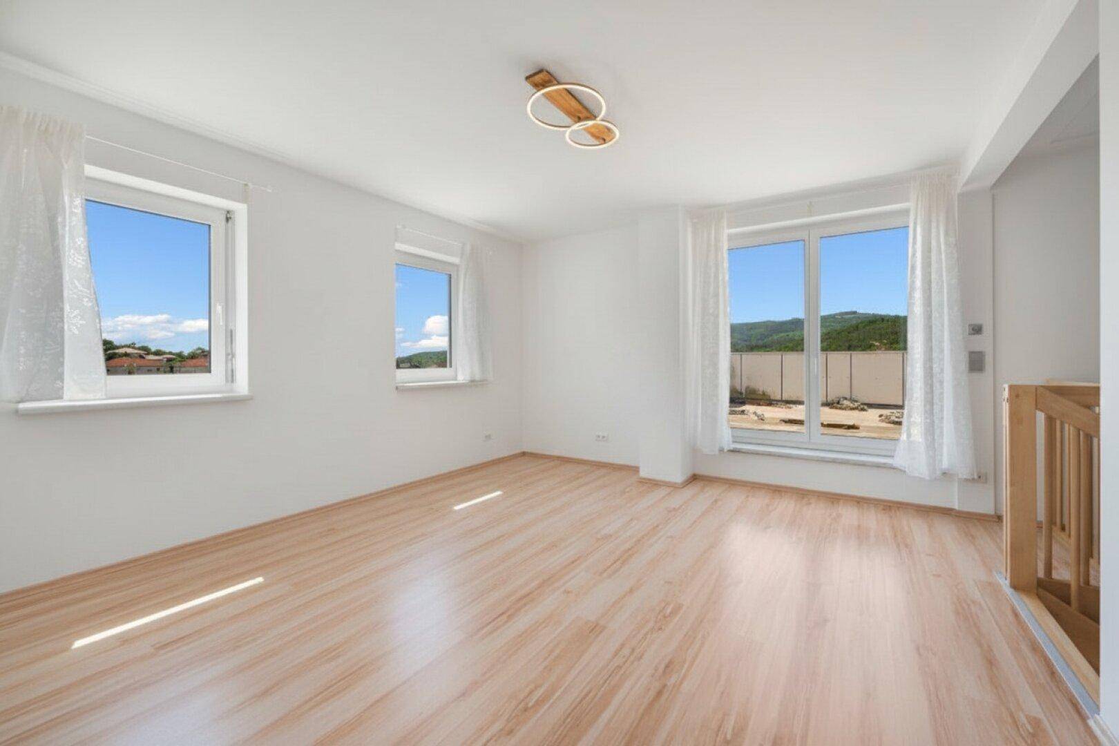 Helles, leeres Zimmer mit Laminatboden, weißen Wänden und Fenstern mit Blick auf die Landschaft.