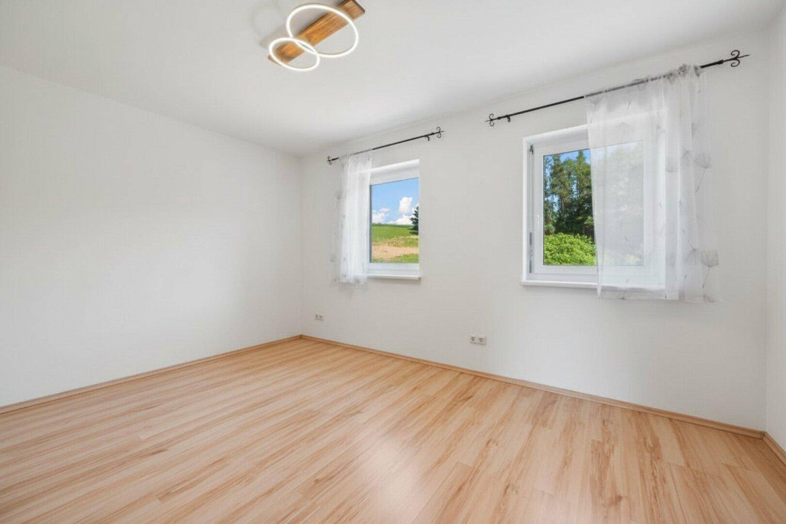Helles, leeres Zimmer mit Laminatboden und zwei Fenstern, die einen Blick ins Grüne bieten.