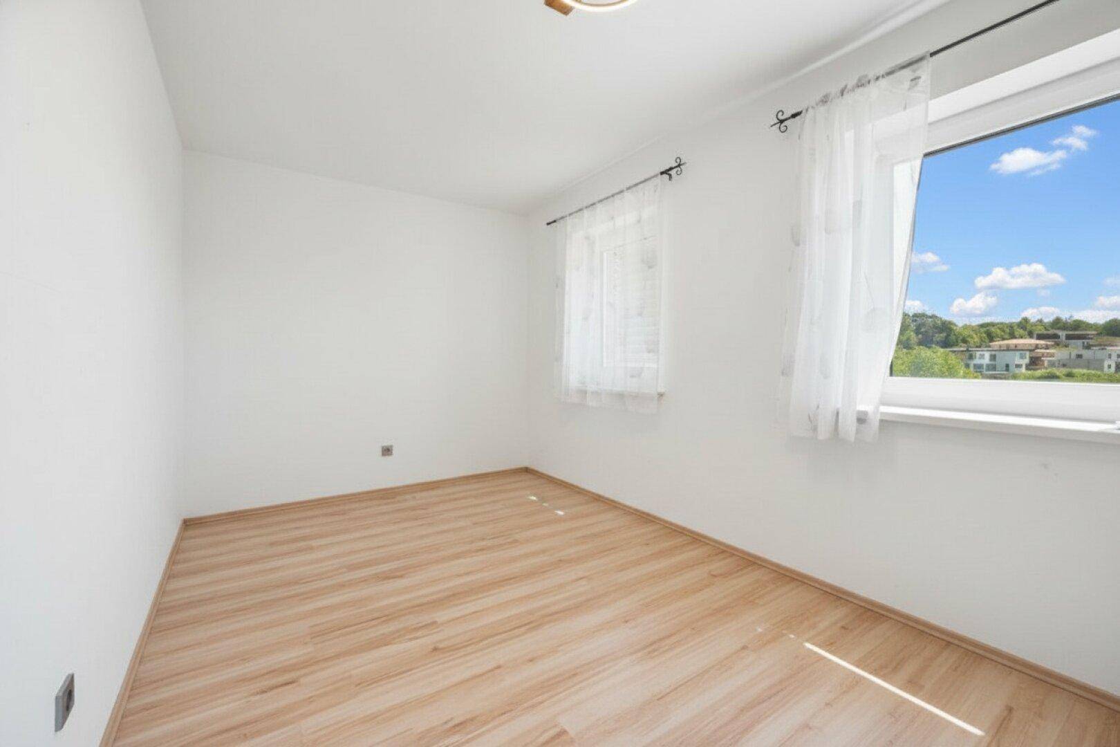 Leeres Zimmer mit hellem Laminatboden und einem großen Fenster mit weitem Ausblick.