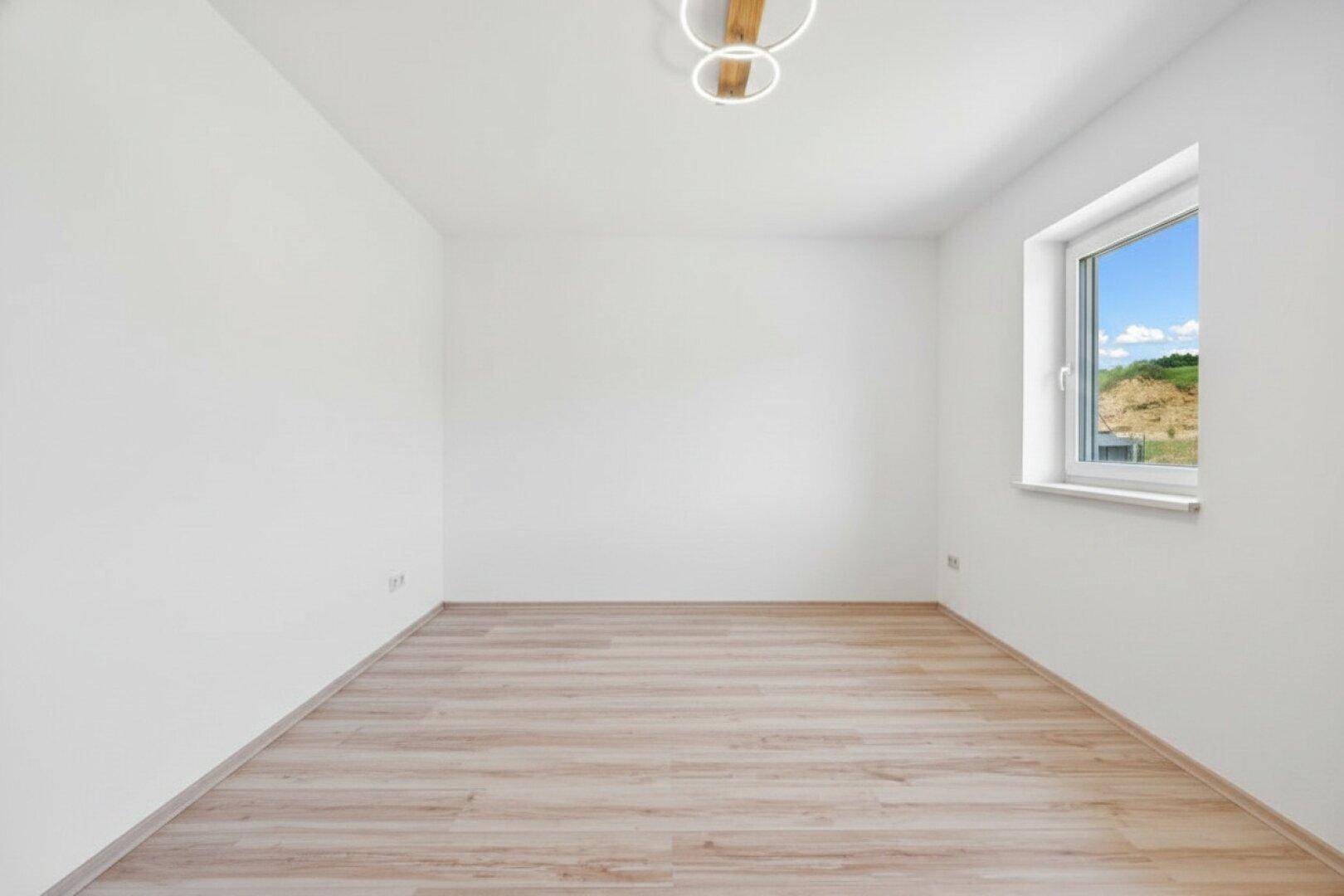 Helles, leeres Zimmer mit Laminatboden und einem Fenster mit Blick auf eine grüne Anhöhe.