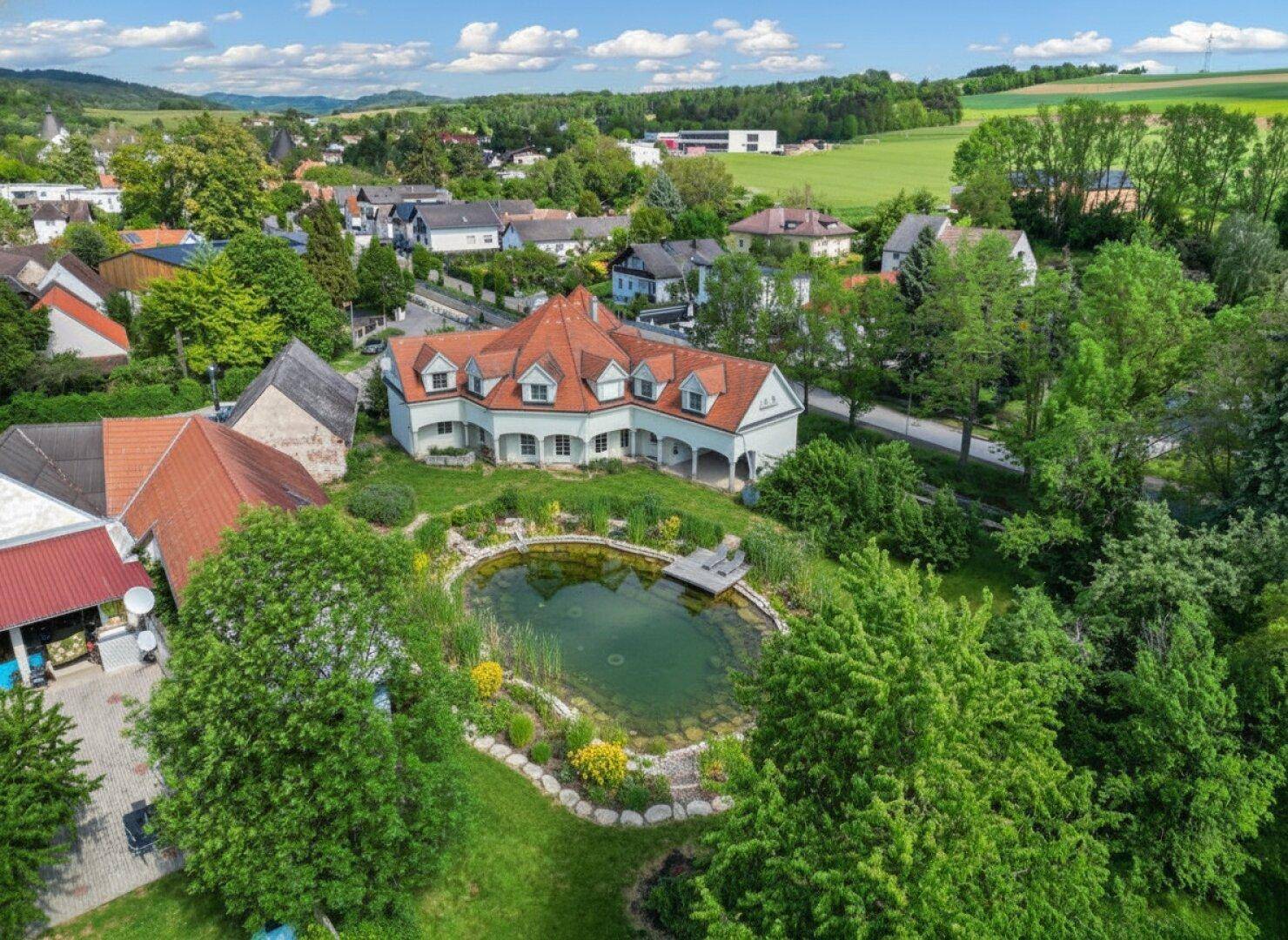 Luftaufnahme eines Anwesens mit großem Haus, natürlichem Teich und weitläufiger grüner Landschaft.