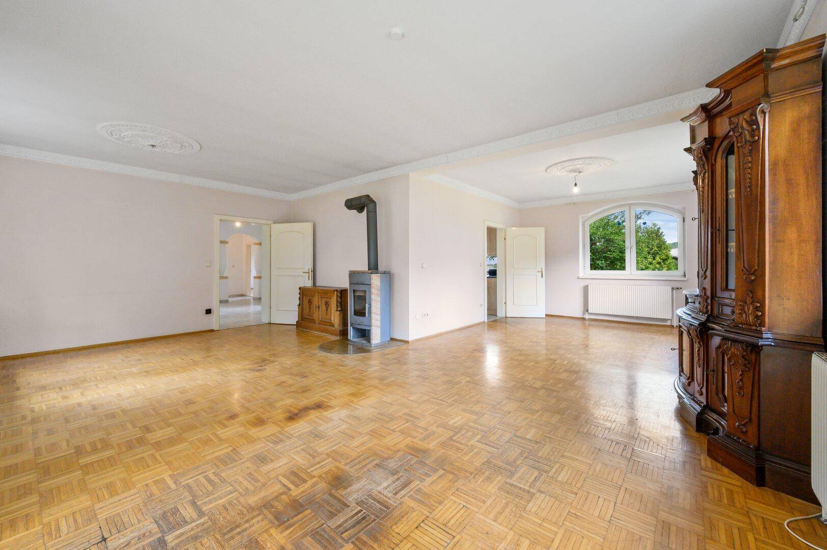 Weitläufiges Wohnzimmer mit Parkettboden, Kaminofen und Zugang zu weiteren Räumen.
