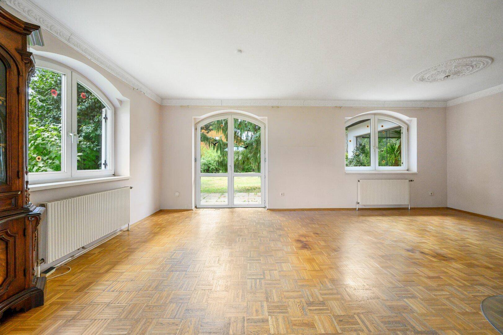 Geräumiges Wohnzimmer mit Fischgrätparkett, hohen Fenstern und Stuckelementen an der Decke.