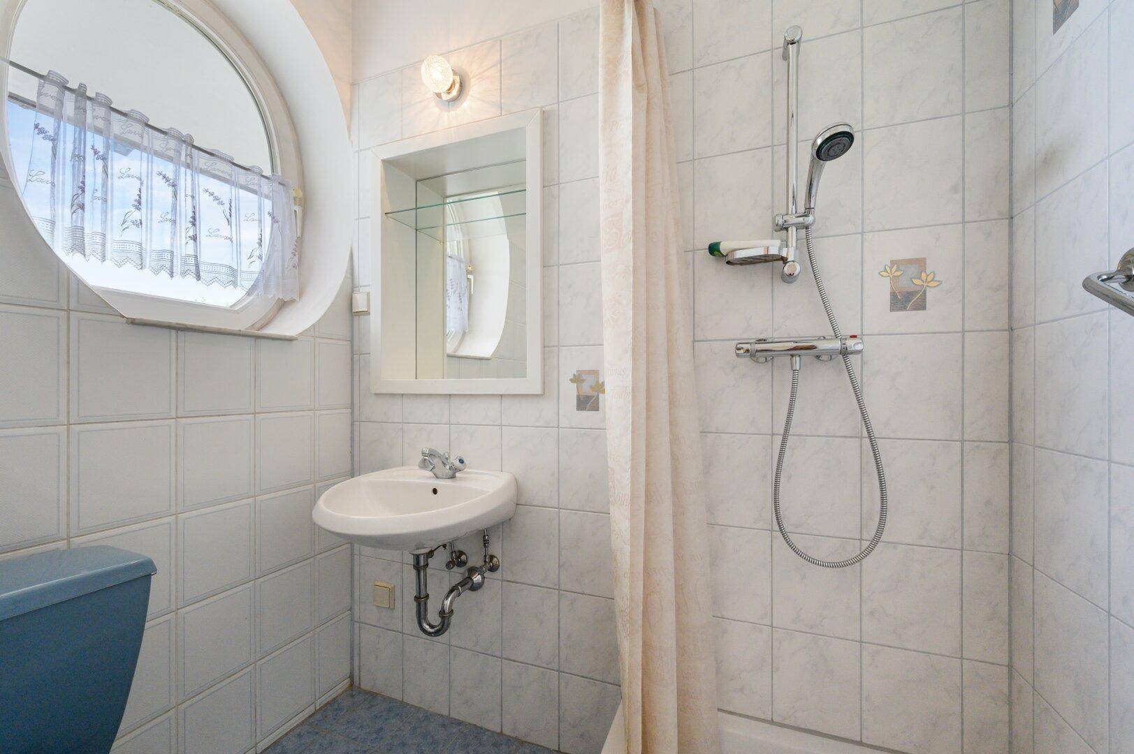 Kleines Badezimmer mit Dusche, Waschbecken und einem runden Fenster.