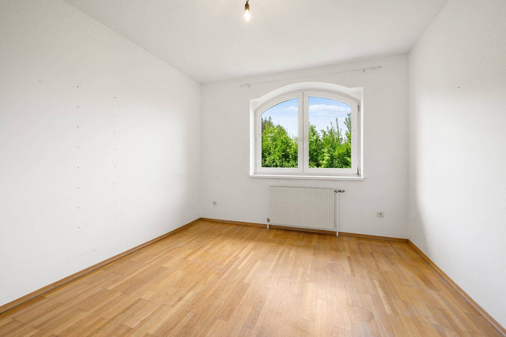 Helles Zimmer mit Holzboden und einem großen Bogenfenster mit Blick ins Grüne.