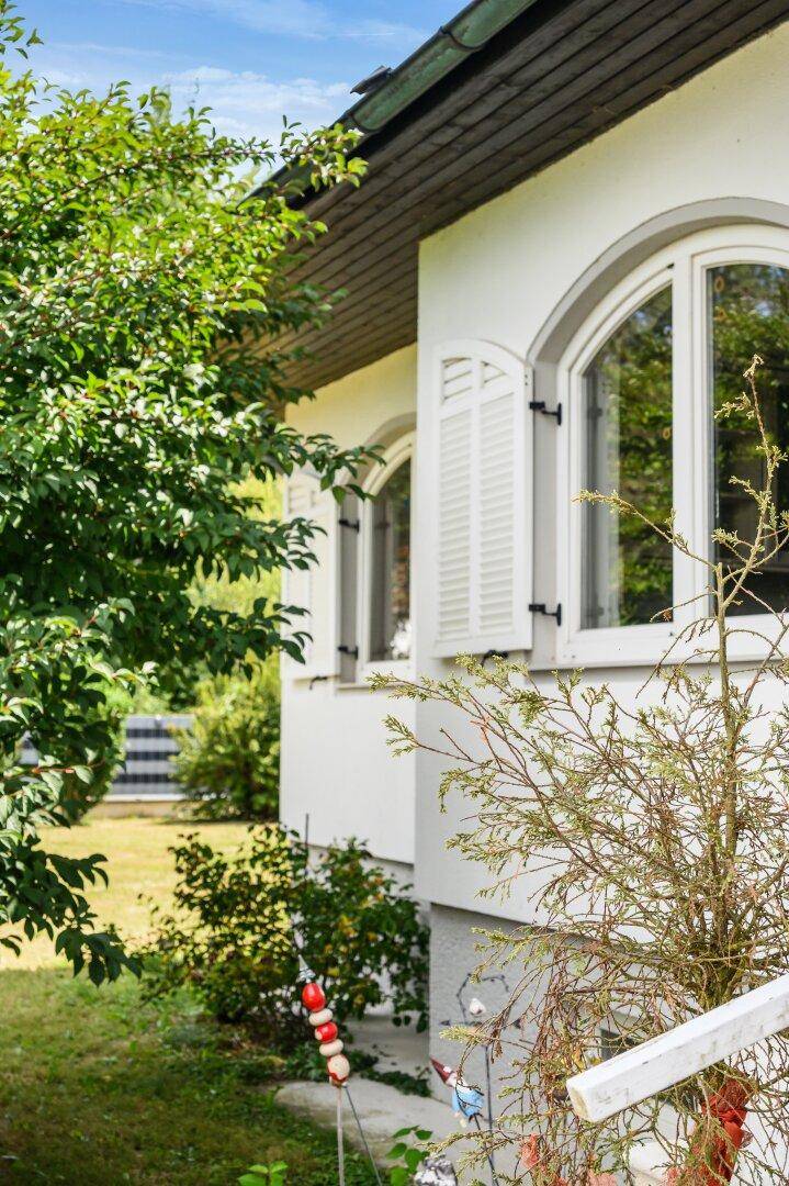 Seitenansicht des Hauses mit Rundbogenfenstern und weißen Fensterläden, umgeben von Grün.