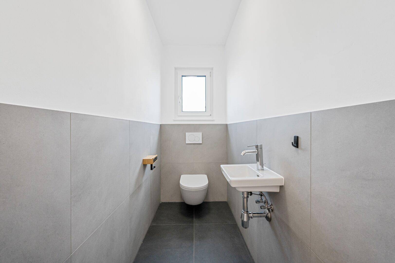 Modernes Gäste-WC mit grauen Fliesen, wandhängendem WC und kleinem Waschbecken.