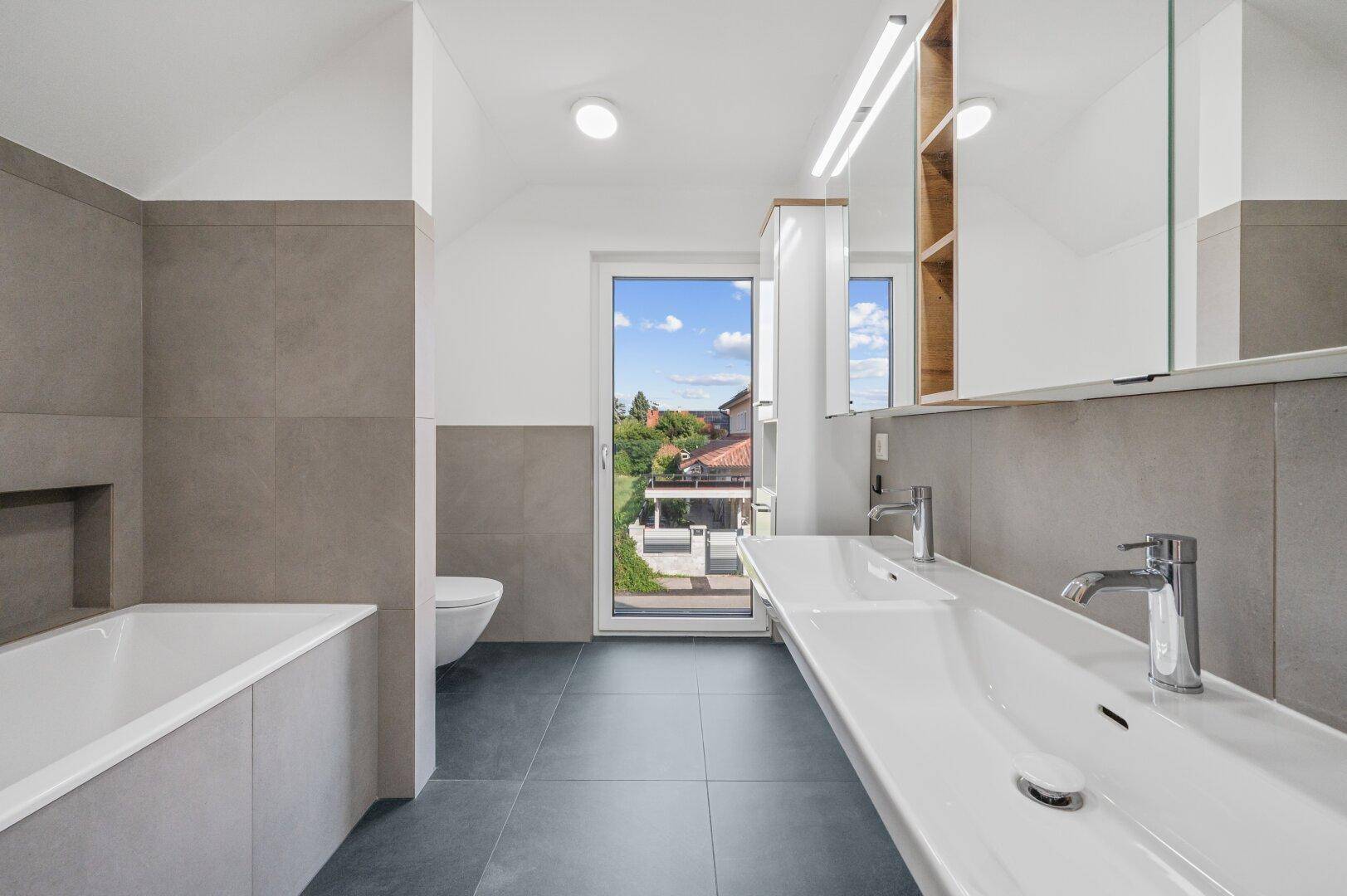 Modernes Badezimmer mit grauen Fliesen, Doppelwaschbecken, Badewanne und Zugang nach draußen.
