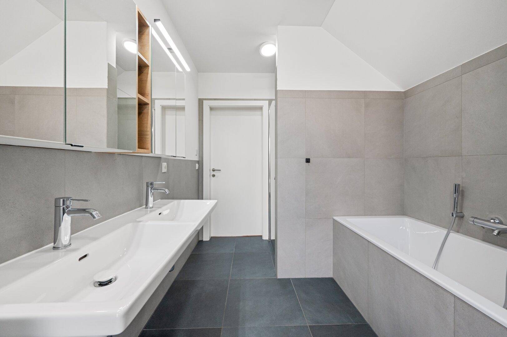 Helles Badezimmer mit grauen Fliesen, Doppelwaschbecken und integrierter Badewanne.
