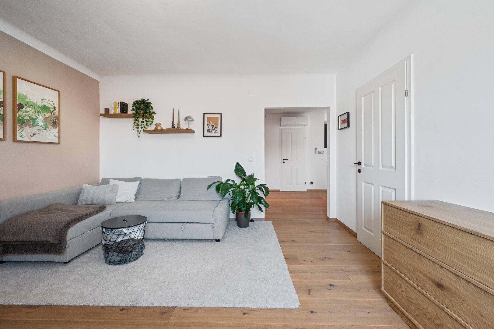 Geräumiges Wohnzimmer mit hellem Sofa, Holzboden und Zugang zu weiteren Räumen.