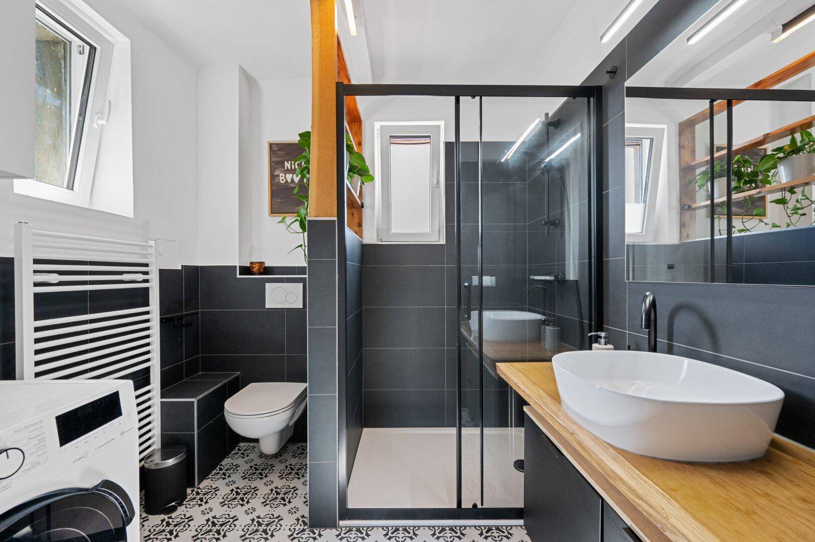 Modernes Badezimmer mit dunklen Fliesen, begehbarer Dusche, Waschbecken und Fenster für Tageslicht.