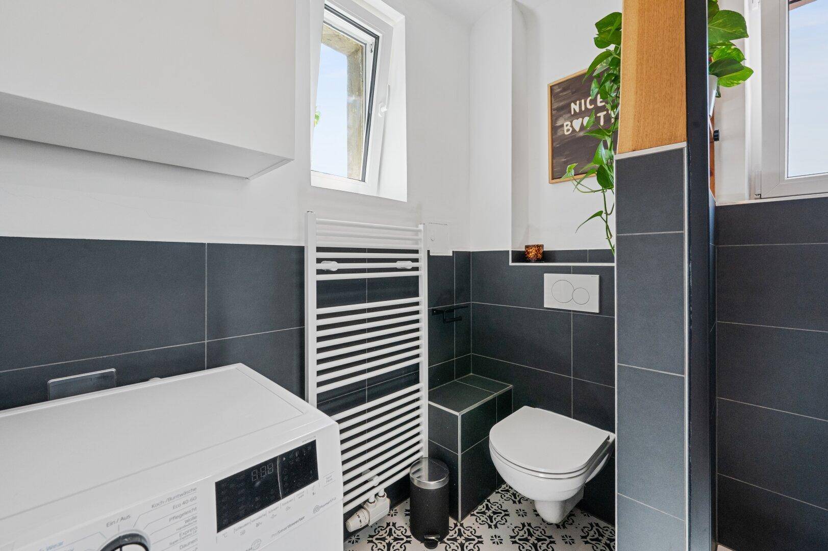 Modernes Badezimmer mit dunklen Fliesen, Wand-WC, Waschmaschine und beheiztem Handtuchhalter.