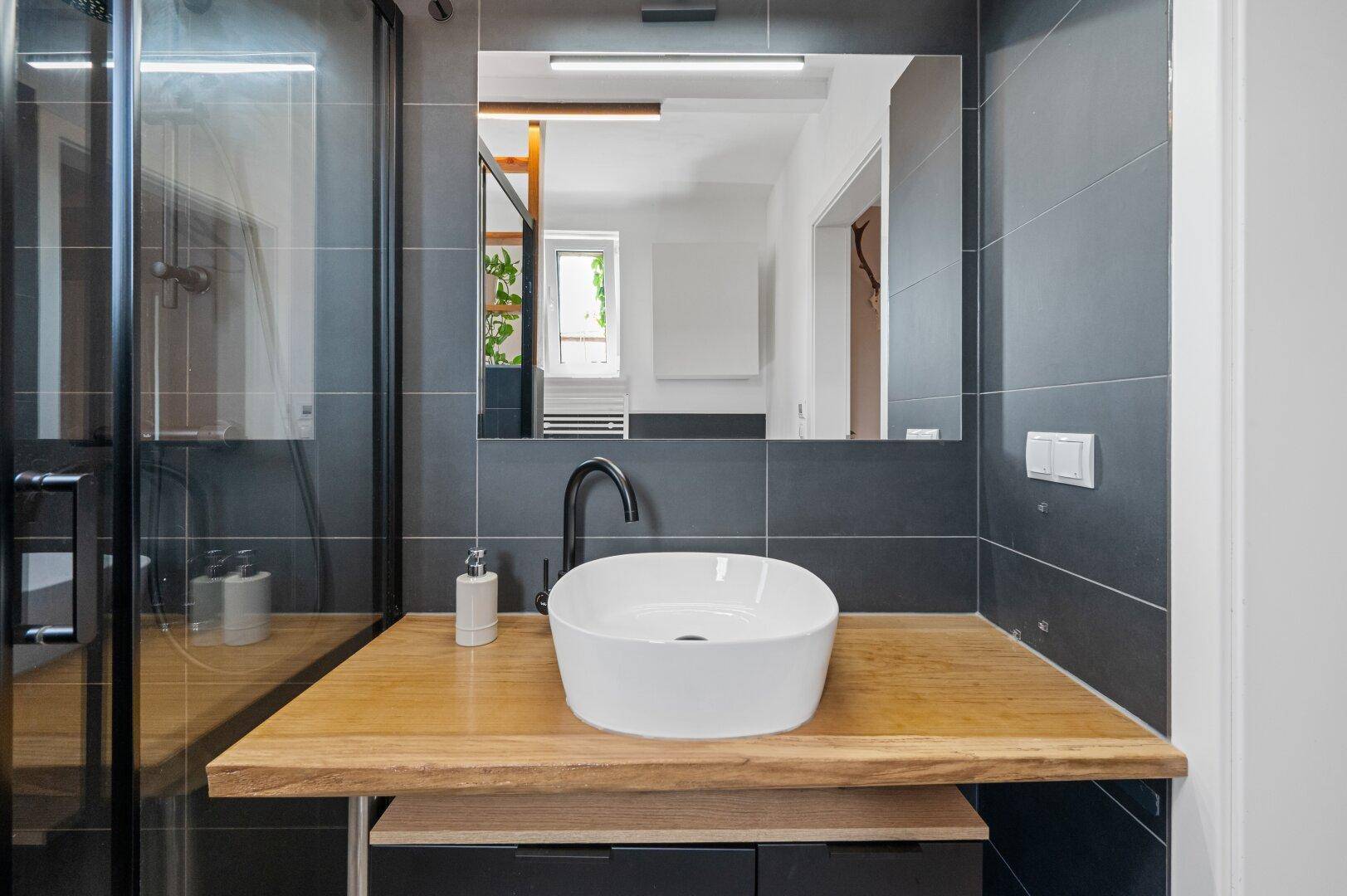 Modernes Waschbecken mit Holzplatte, schwarzem Wasserhahn und großem Spiegel im Badezimmer.