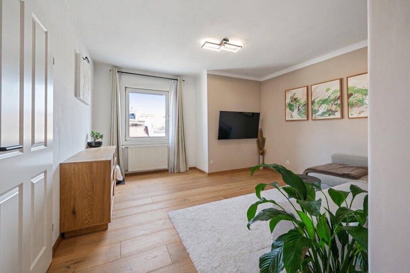 Helles Wohnzimmer mit Holzboden, Teppich, Fernseher und Blick auf die Stadt durch das Fenster.