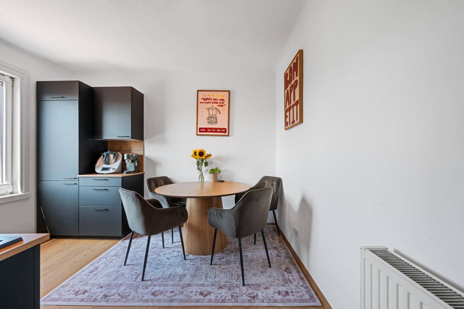 Heller Essbereich in der Küche mit Holztisch, modernen Stühlen und dunklen Küchenschränken.