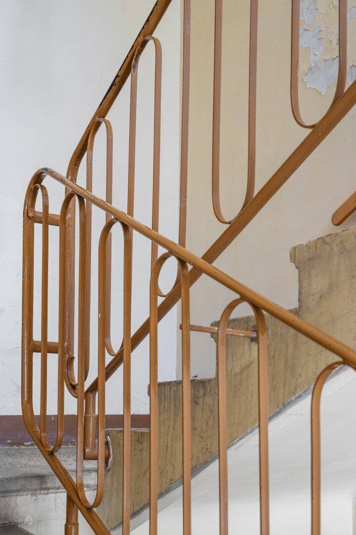 Altes Treppenhaus mit charakteristischem braunem Metallgeländer und sichtbaren Gebrauchsspuren an den Wänden.