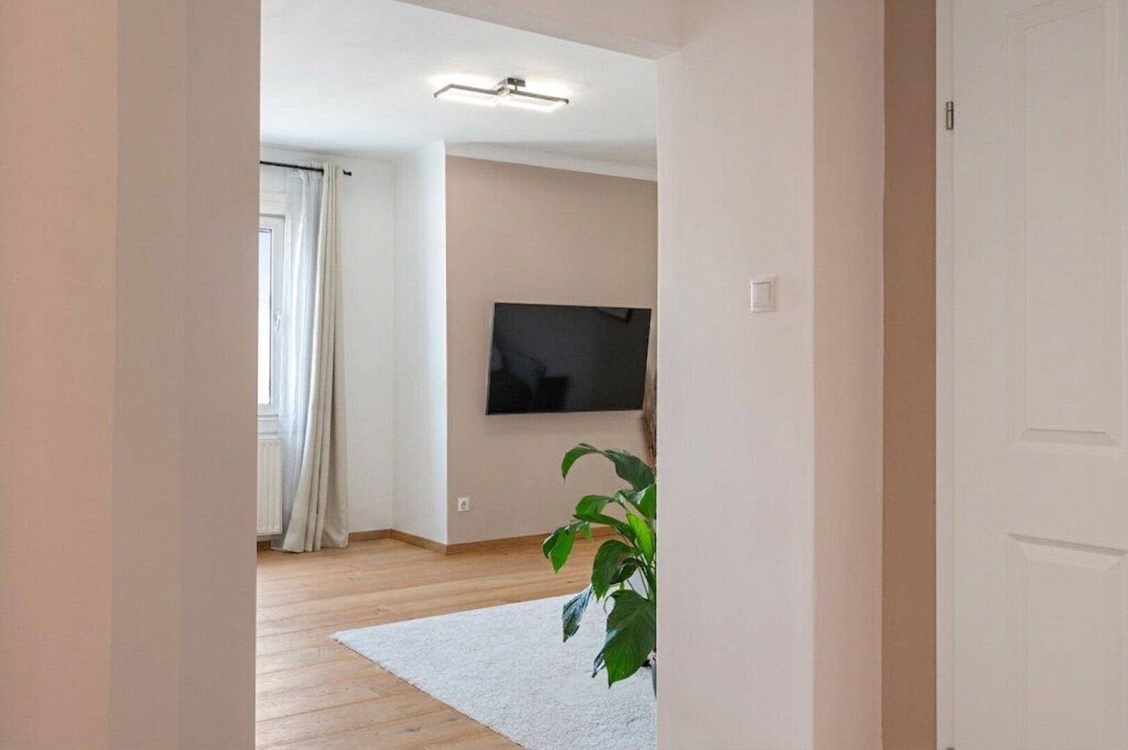 Blick vom Flur in das helle Wohnzimmer mit Holzboden und einem an der Wand montierten Fernseher.